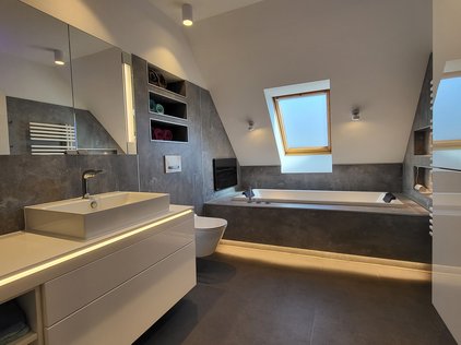 Man schaut in ein modernes Badezimmer. Links ein großer heller Waschtisch, mit aufgestelltem Waschbecken. Darüber ein Spiegelschrank. Hinter dem Waschtisch blickt man auf eine Toilette. Rechts daneben erkennt man eine in einen grauen Steinblock eingelassene weiße Badewanne. Rechts schaut man auf einen großen Badezimmerschrank mit Hochglanzfronten.