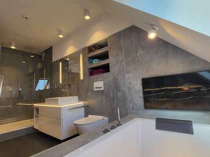 Man sieht einen Ausschnitt eines modernen Badezimmers. Links sieht man eine moderne Dusche, komplett mit Glaswänden. Rechts daneben befindet sich ein weißer Waschtisch, wobei das Becken auf der Waschtischplatte steht und nicht eingelassen ist. An der Wand dahinter ist ein großflächiger Spiegelschrank zu erkennen. Rechts daneben erkennt man ein freihängendes Wand-WC in weiß.